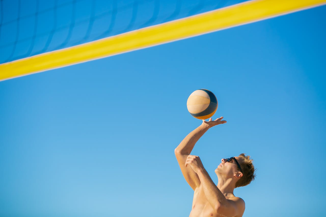 A man in sunglasses serves a volleyball outdoors on a sunny day.